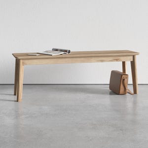May include: A light brown wooden bench with four tapered legs and a rectangular seat. An open magazine rests on the top surface of the bench, and a tan leather satchel bag is positioned on the floor leaning against one of the bench legs.