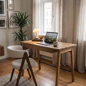 May include: A wooden desk with a laptop displaying the word created, a small lamp, and a mug. Beside the desk is a chair with a white textured fabric seat and backrest, supported by a dark wooden frame with angled legs.