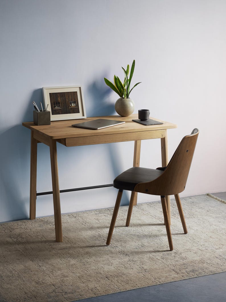 May include: A light oak wood desk with a single drawer and a thin black metal support bar, paired with a matching wood chair featuring a dark brown upholstered seat. On the desk sits a laptop, a framed picture, a plant, and a small cup.