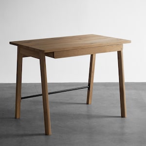 May include: A rectangular wooden desk featuring a light brown oak grain finish, a single integrated drawer positioned beneath the tabletop, four angled legs, and a thin black metal support bar connecting the two rear legs.