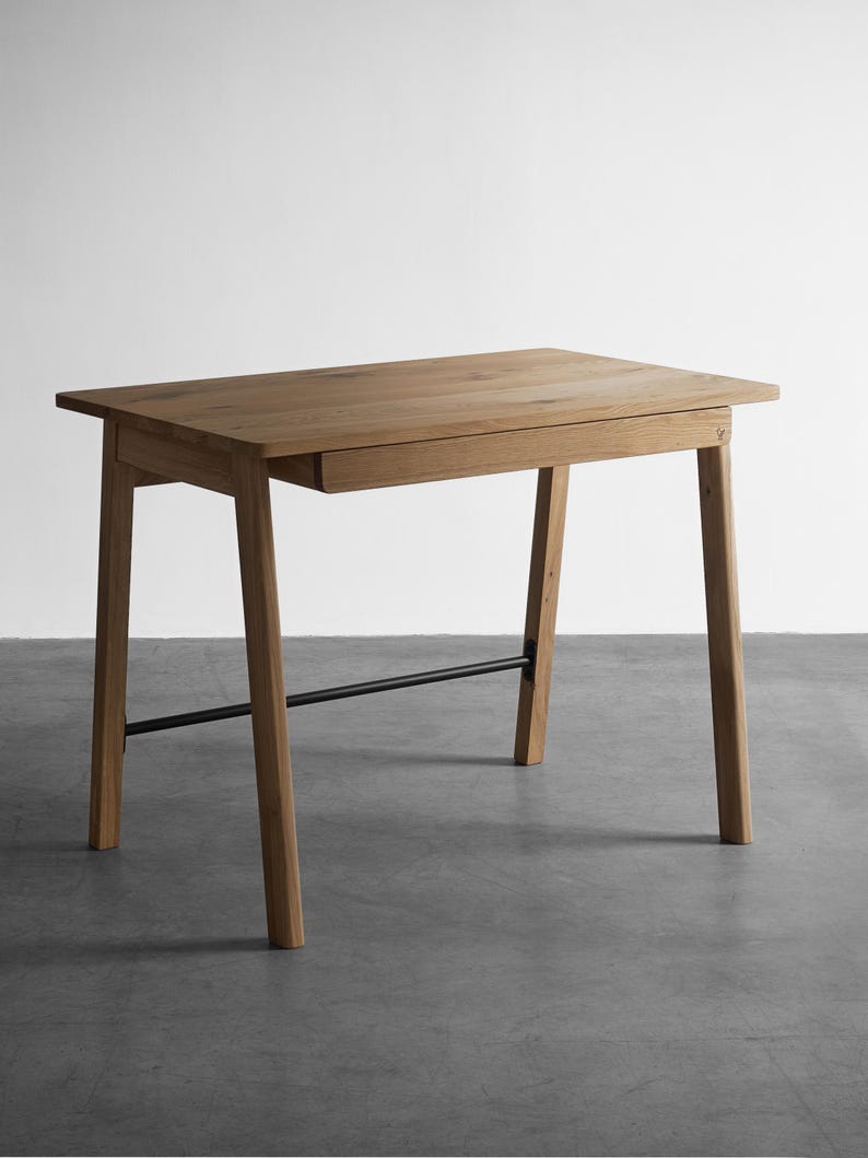 May include: A rectangular wooden desk featuring a light brown oak grain finish, a single integrated drawer positioned beneath the tabletop, four angled legs, and a thin black metal support bar connecting the two rear legs.