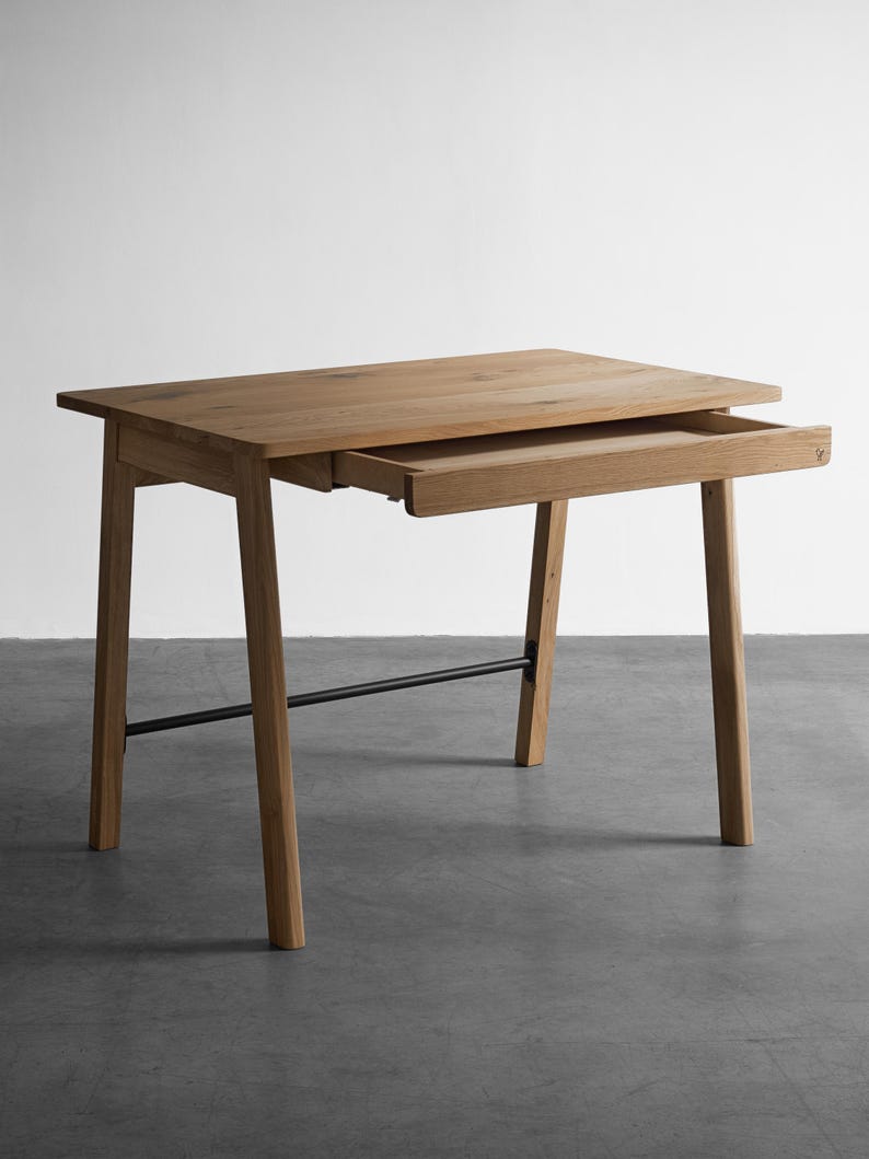 May include: A light brown wooden desk with a rectangular top and a single pull-out drawer. The desk features four angled legs connected by a thin black horizontal metal support bar positioned between the two rear legs.