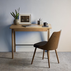 May include: A light wood desk with a single drawer and a black metal support bar, paired with a matching wood chair featuring a dark seat. On the desk sits a silver laptop, a potted green plant, a framed picture, and a gray pen holder.