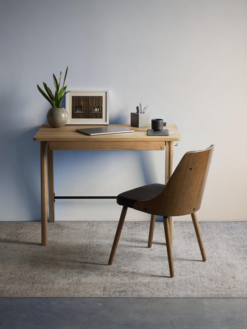 May include: A light wood desk with a single drawer and a black metal support bar, paired with a matching wood chair featuring a dark seat. On the desk sits a silver laptop, a potted green plant, a framed picture, and a gray pen holder.
