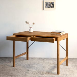 Peut inclure: Un bureau rectangulaire en bois au fini brun naturel, doté de deux tiroirs coulissants à l'avant, de quatre pieds robustes et de fines entretoises de support en métal noir. Un vase blanc avec des fleurs séchées et une pile de livres sont posés.