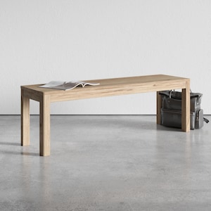 May include: A light-colored wooden bench with a rectangular top and four sturdy legs. An open magazine rests on the bench's surface. A dark gray backpack leans against the bench's side.