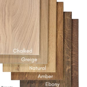 May include: A collection of six rectangular oak wood samples arranged in a cascading stack. The finishes are labeled as Chalked, Greige, Natural, Amber, Ebony, and Walnut. The text WOODWERK appears in the top left corner with a bird icon.