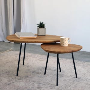 Könnte beinhalten: Zwei Holz-Beistelltische mit schwarzen Metallbeinen. Die Tische haben eine ovale Form und eine natürliche Holzoberfläche. Eine kleine Topfpflanze, ein Stapel Bücher und eine Tasse mit gepunktetem Muster befinden sich auf den Tischen. Die Tische stehen auf einem gemusterten Teppich.