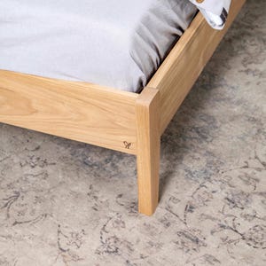 May include: Close-up of a wooden bed frame with a light gray sheet on top. The bed frame is made of natural wood and has a simple design. A small bird logo is visible on the side of the frame.