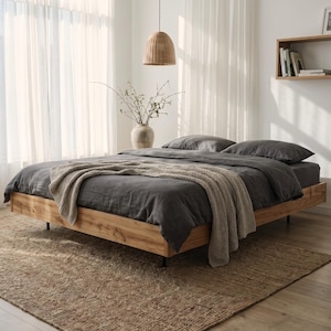 May include: A low-profile wooden bed frame with a natural grain finish, featuring dark charcoal grey bedding and a textured beige knit throw blanket draped across the foot. The bed is positioned on a large, woven jute area rug in a bright room.