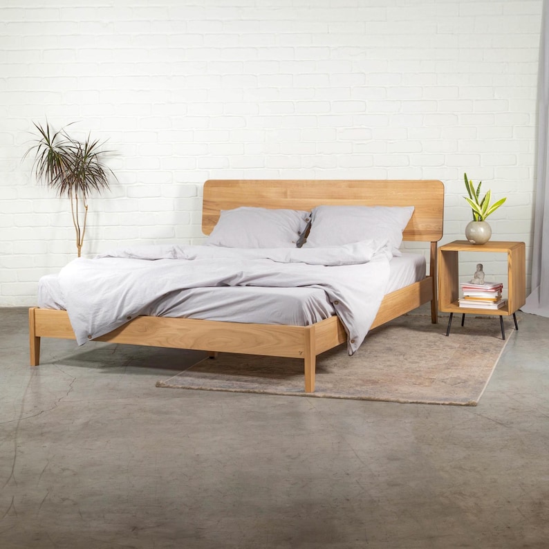 May include: A wooden bed frame with a light wood finish and a headboard with rounded edges. The bed is made up with a white duvet and two white pillows. A small wooden nightstand with black legs is next to the bed.