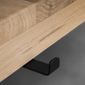 May include: A light-colored wooden shelf featuring a visible grain pattern and a smooth, flat surface. Attached to the underside of the wooden board is a single, matte black metal hook with a curved, upturned end for hanging items.