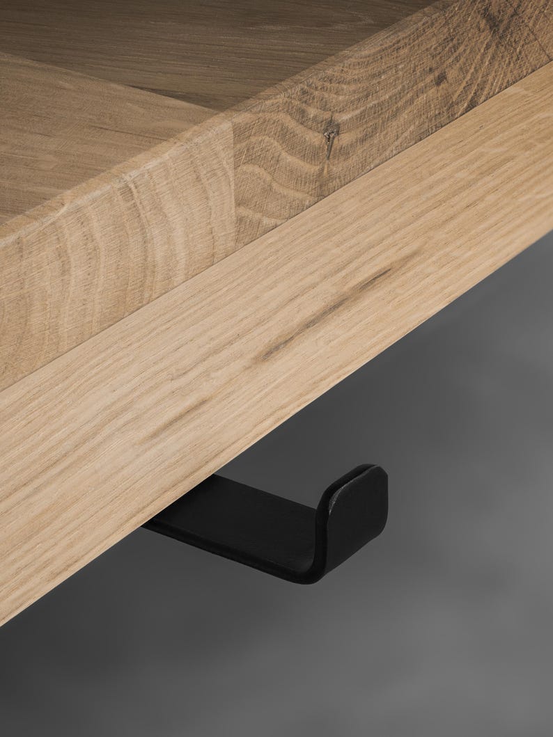 May include: A light-colored wooden shelf featuring a visible grain pattern and a smooth, flat surface. Attached to the underside of the wooden board is a single, matte black metal hook with a curved, upturned end for hanging items.