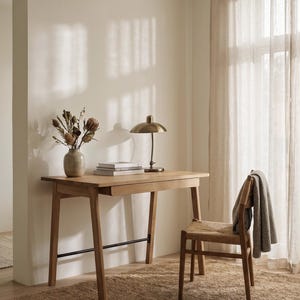 May include: A light-toned wooden desk with a single drawer and a black metal support bar, accompanied by a wooden chair with a woven seat. On the desk sits a brass-colored metal lamp, a stack of books, and a vase containing dried flowers.