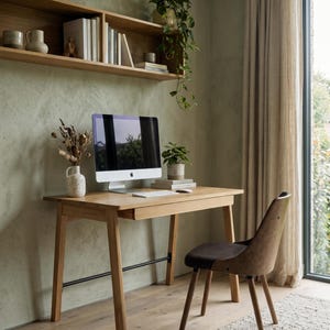 May include: A light oak wood desk with a single drawer and a black metal support bar, paired with a brown upholstered chair. On the desk sits an all-in-one computer, a white keyboard, a mouse, a potted plant, and a vase with dried flowers.