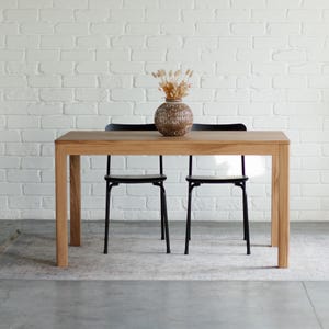 Puede incluir: Una mesa de comedor de madera clara con una parte superior rectangular y cuatro patas resistentes. Dos sillas de metal negro están colocadas debajo de la mesa. Un jarrón decorativo con flores secas está sobre la mesa. El fondo es una pared de ladrillos blancos.