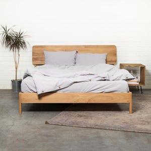 May include: A wooden bed frame with a light gray duvet and two pillows. The bed frame has a headboard with a curved top and a footboard with straight lines. The bed is sitting on a gray concrete floor with a rug in front of it.