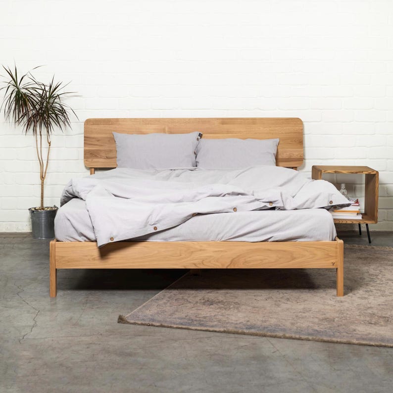 May include: A wooden bed frame with a light gray duvet and two pillows. The bed frame has a headboard with a curved top and a footboard with straight lines. The bed is sitting on a gray concrete floor with a rug in front of it.
