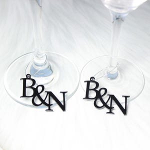 以下が含まれることがあります： 2つのワイングラスのステムに、"B&N"の文字が書かれた黒いガラス製のワインチャームがぶら下がっています。