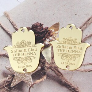 May include: Two gold-coloured, mirror-finish hamsa-shaped acrylic tags featuring an engraved mandala design. The text reads Shilat & Elad THE HENNA 14.09.2025. Each tag includes a small circular hole at the top and bottom for attachment purposes.