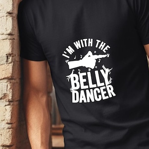 Puede incluir: Camiseta azul marino con el texto blanco "I'm with the Belly Dancer". El gráfico presenta la silueta de una bailarina de la danza del vientre. La camiseta es de cuello redondo clásico.