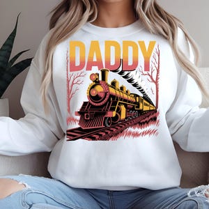Puede incluir: Sudadera blanca con un diseño gráfico de un tren antiguo y la palabra "DADDY" en naranja y rosa. El tren es negro y amarillo con detalles rojos, y sale humo de la chimenea. El diseño está sobre un fondo blanco.