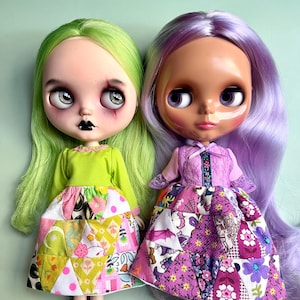 Puede incluir: Dos muñecas Blythe con ojos grandes y pelo largo. Una muñeca tiene el pelo verde, una blusa verde lima y una falda patchwork con triángulos rosas y amarillos. La otra muñeca tiene el pelo morado, una blusa lavanda y una falda patchwork con estampados morados y rosas.
