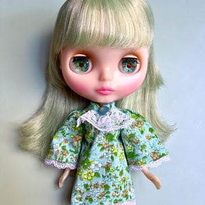 May include: A doll with green hair and large, expressive eyes. The doll wears a light blue dress with a floral pattern in yellow, orange, and white. The dress has white lace trim at the collar and sleeves.
