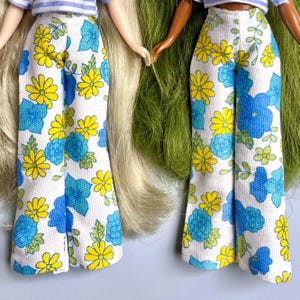 May include: Two doll outfits featuring white, wide-leg pants with a floral pattern of yellow and blue flowers. The dolls wear striped tops. The outfits are reminiscent of the 1970s fashion.