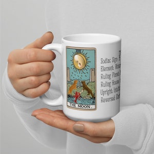 May include: White ceramic mug with a colorful tarot card design featuring The Moon card from the Rider-Waite deck. The card depicts a crescent moon, two towers, a dog, a crab, and a path. The text "XVIII THE MOON." is printed below the card. The text "Zodiac Sign: Pisces, Element: Water, Ruling Planet: Jupiter, Ruling House: 12th, Upright: Intuition, Reversed: Clarity" is printed to the right of the card.