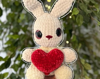 Op de afbeelding: Een witte gehaakte bunny die een rood, hartvormig gehaakt kussen vasthoudt. De bunny heeft grote zwarte ogen en een rode neus.