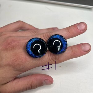 以下が含まれることがあります： 黒い虹彩と白い三日月型の瞳を持つ、丸いガラスのような目。目の外側は、銀河のような模様の深い青色です。「#1」の数字が紫色のインクで書かれています。