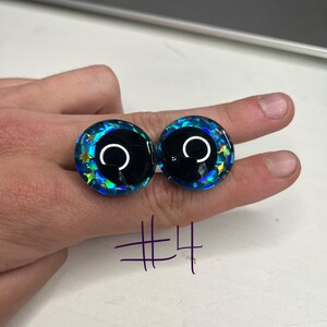 以下が含まれることがあります： 黒い中心と白い三日月のある、ガラスのような丸いカボションが2つ。外側の縁は鮮やかな青色で、虹色のグリッターと星型のアクセントが付いています。「#4」の数字が紫でカボションの下に書かれています。