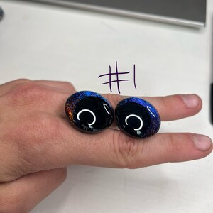 以下が含まれることがあります： 青、オレンジ、金色の銀河のようなデザインの、2つの楕円形の黒いカボションストーン。各石には白い三日月型のアクセントがあります。番号「#1」が石の上に書かれています。
