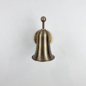 May include: A brass wall sconce featuring a bell-shaped shade and a circular backplate. The sconce has a warm, metallic finish, typical of brass. The design incorporates a small, spherical detail at the top, adding a touch of elegance to the lighting fixture.
