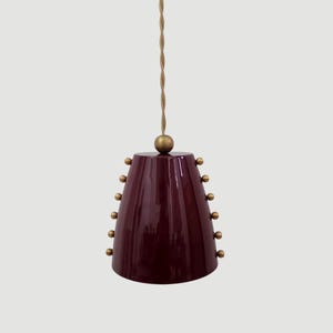 May include: A pendant light fixture with a deep burgundy shade and brass accents. The shade is cone-shaped with a series of small brass spheres around the bottom. A gold-colored cord suspends the light from above.