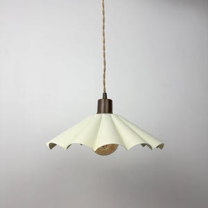 May include: Cream-coloured pendant light with a scalloped shade. The light fixture has a bronze-coloured base and a twisted beige cord. The bulb is visible beneath the shade. The light hangs from the ceiling against a plain white background.