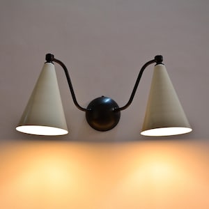 Può includere: Applique da parete doppio con struttura nera e base circolare. Presenta due paralumi conici color crema. La luce calda crea un bagliore soffuso sulla parete.