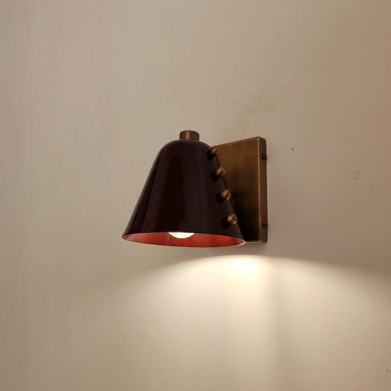 Handcrafted Wall Sconce with A Rich Wine Colored Tapered Cone Shade, Solid Brass Construction, and Decorative Rivet Details, Shown in A Compact Profile that Suits Mid Century Inspired Interior Lighting, Frost Lights.