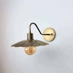 Peut inclure: Applique murale en laiton avec un abat-jour festonné et un bras incurvé. L'ampoule est visible à l'intérieur de l'abat-jour. Le luminaire est monté sur une base ronde en laiton. Le design est d'inspiration vintage.