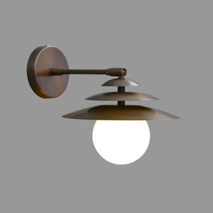 Peut inclure: Une applique murale composée d'une plaque de fixation circulaire couleur bronze et d'un bras horizontal supportant un abat-jour à étages. L'abat-jour présente trois disques métalliques superposés en bronze foncé et une sphère en verre blanc.