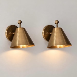 May include: A pair of wall-mounted sconces featuring a conical brass shade and a circular backplate. Each light fixture has a small spherical finial at the top of the shade, with a warm light emitting from the open bottom of the metal cone.