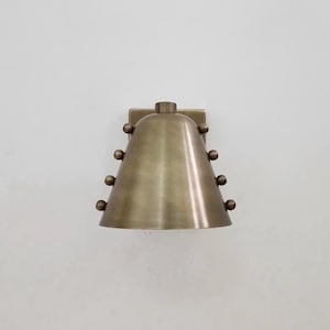 May include: A brass-coloured wall sconce with a conical shade and a square mounting plate. The sconce has spherical accents along the shade's sides. The fixture is designed for wall mounting and provides ambient lighting.