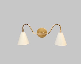 May include: A double-light wall sconce with a brass finish. The fixture features two curved arms, each supporting a white, cone-shaped shade. The sconce is mounted on a circular backplate.
