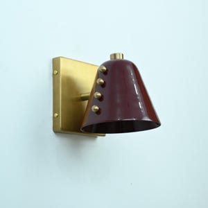 A Refined Wine Shade Wall Sconce with Brass Construction and Rivet Accents Is Shown from An Angled Perspective, Emphasizing the Cone Shape and Compact Architectural Silhouette of The Fixture, Frost Lights.
