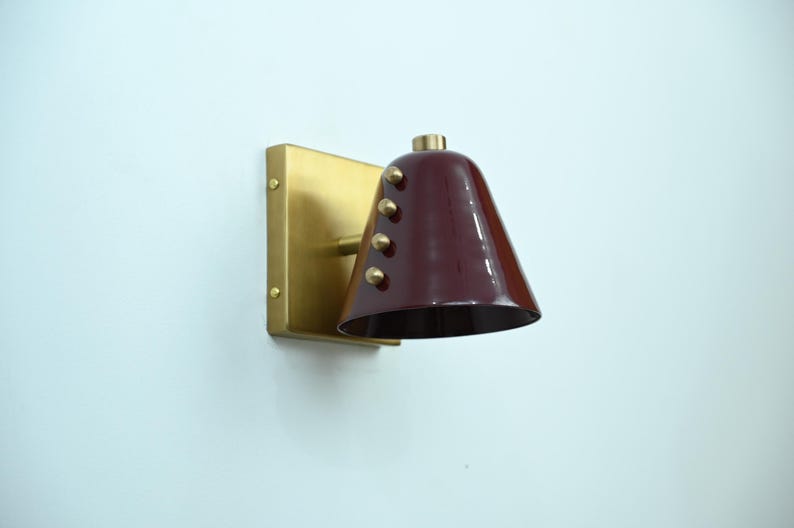 A Refined Wine Shade Wall Sconce with Brass Construction and Rivet Accents Is Shown from An Angled Perspective, Emphasizing the Cone Shape and Compact Architectural Silhouette of The Fixture, Frost Lights.