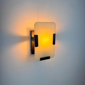 May include: A rectangular wall sconce featuring a matte black metal frame and a translucent, off-white glass panel. The fixture emits a warm, golden-yellow light that illuminates the surrounding area, creating a soft glow against the wall surface.