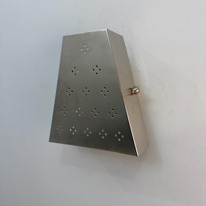 May include: A brushed metal wall sconce with a trapezoidal shape. The sconce has a series of small, symmetrical holes on the front and a small, round knob on the side. The sconce is mounted on a white wall.