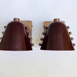 Op de afbeelding: Twee bijpassende wandlampen met bordeauxrode, klokvormige kappen en messing accenten. Elke lamp heeft een rechthoekige messing achterplaat en een reeks kleine, ronde messing versieringen. De lampen zijn aan een witte muur bevestigd.