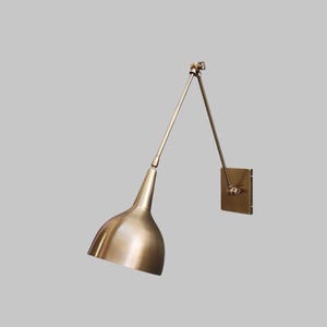 Peut inclure: Une lampe murale en laiton avec un bras réglable et un abat-jour conique. La lampe est fixée à une plaque murale carrée. Le design est minimaliste et moderne, adapté à divers styles d'intérieur. Mesures en centimètres.